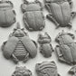 White clay castings on a white background from Iron Orchid Designs Specimens mould. The image shows intricately designed beetles and bugs