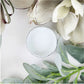 A small round glass jar filled with white paint, photographed from above on a white surface with decorative white flowers and green leaves around it. Paint Couture