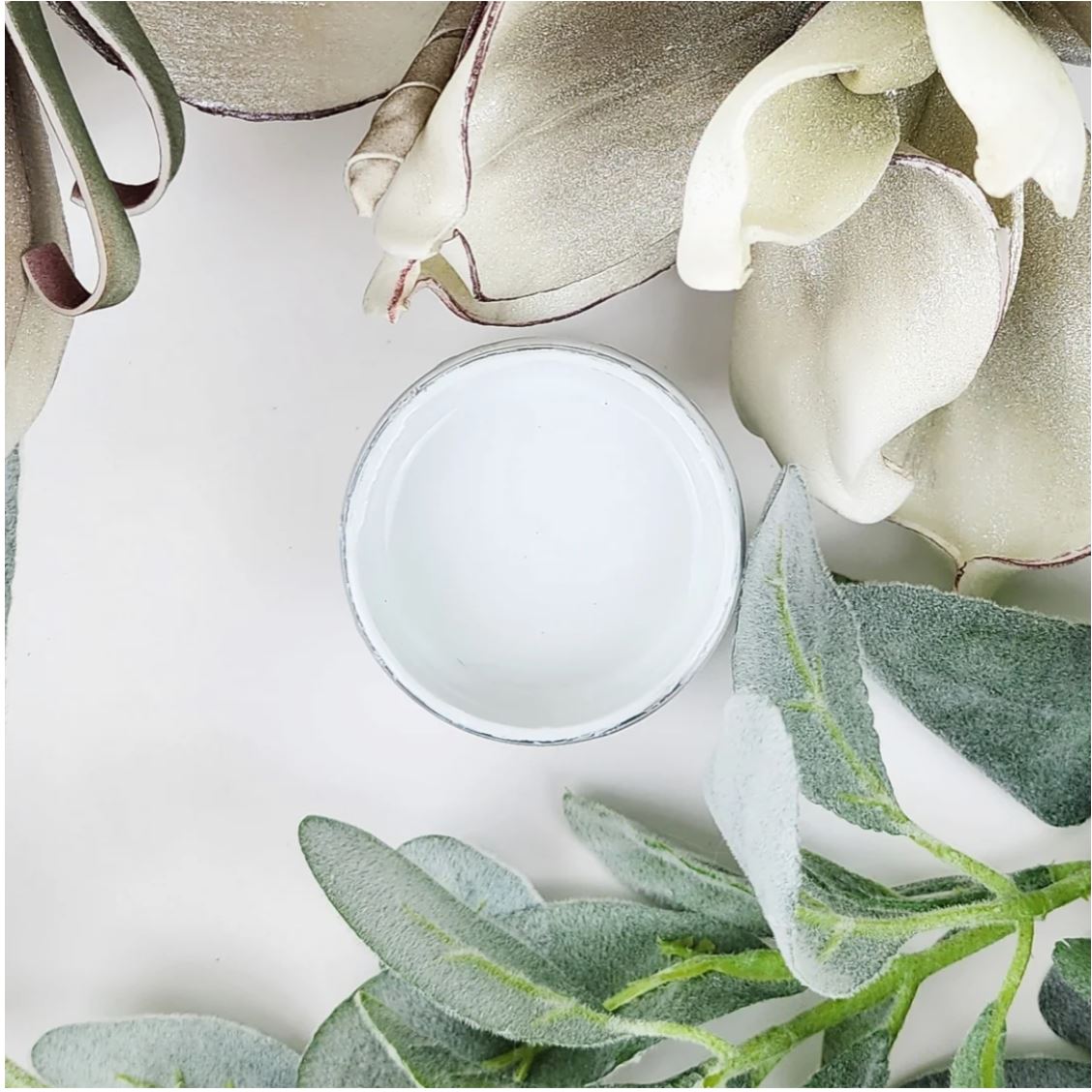 A small round glass jar filled with white paint, photographed from above on a white surface with decorative white flowers and green leaves around it. Paint Couture
