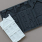 A dark gray silicone mold with embossed house-shaped cavities, shown with a white molded house sample piece on a plain taupe background. From Iron Orchid Designs.