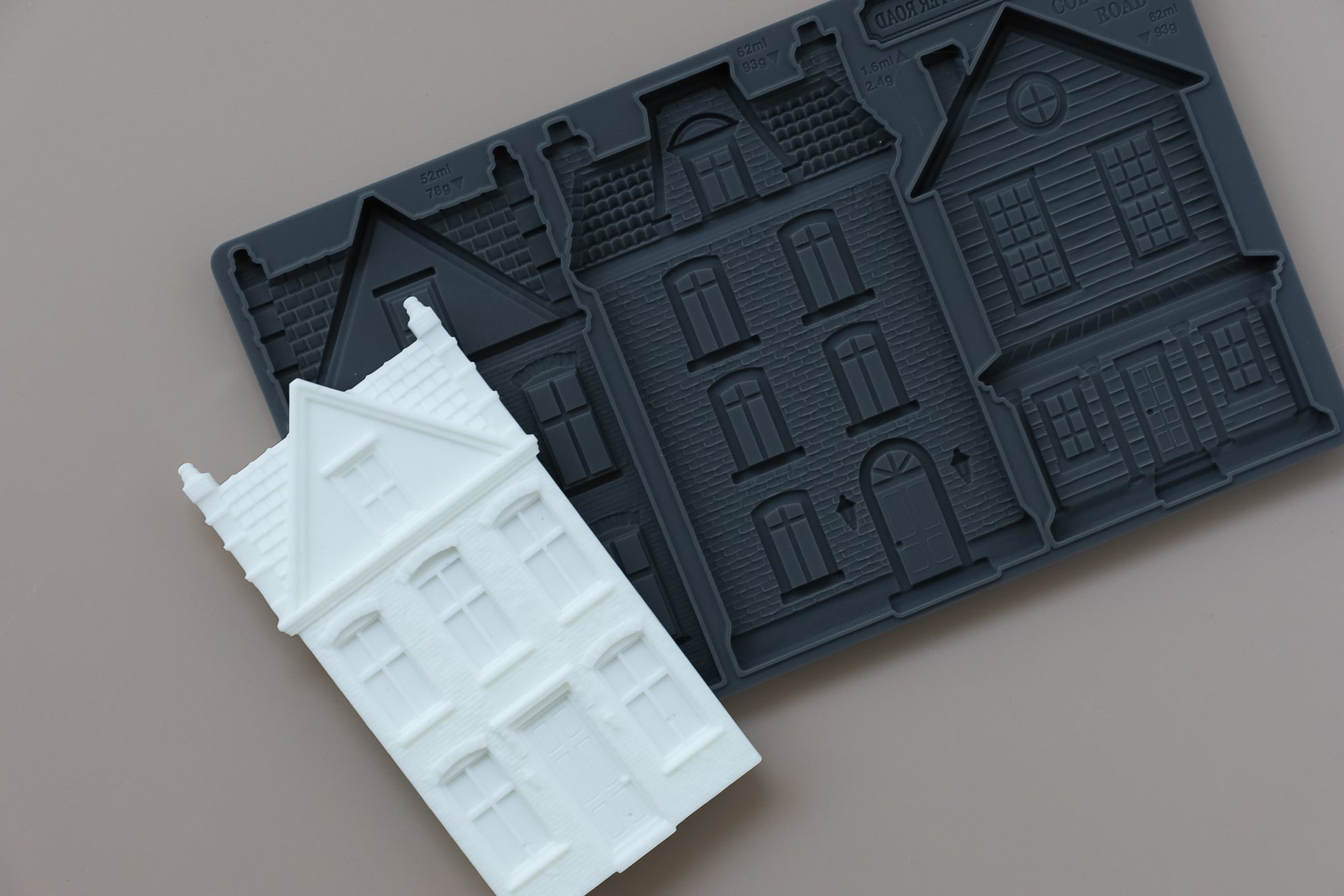 A dark gray silicone mold with embossed house-shaped cavities, shown with a white molded house sample piece on a plain taupe background. From Iron Orchid Designs.