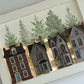 A framed miniature art piece depicting four detailed vintage houses in warm brown, gray, and red tones, set against a background of green pine trees, all enclosed in a white textured frame. From Iron Orchid Designs.