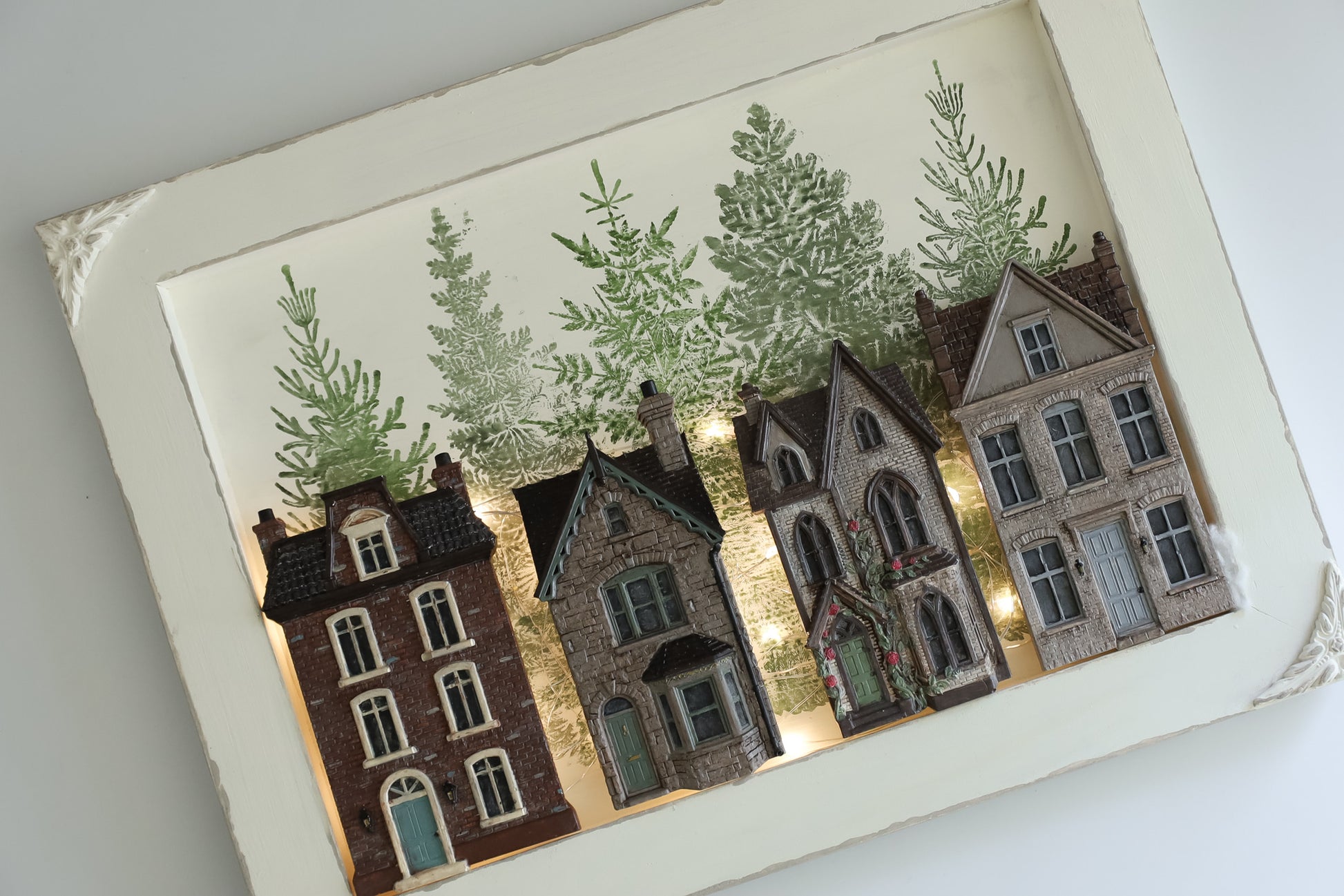 A framed miniature art piece depicting four detailed vintage houses in warm brown, gray, and red tones, set against a background of green pine trees, all enclosed in a white textured frame. From Iron Orchid Designs.