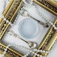 A decorative product shot featuring two ornate silver spoons with shell-shaped handles, a small round container of pale grey paint, and strands of white pearl beads arranged inside an ornate gold picture frame. Paint Couture