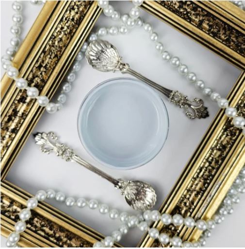 A decorative product shot featuring two ornate silver spoons with shell-shaped handles, a small round container of pale grey paint, and strands of white pearl beads arranged inside an ornate gold picture frame. Paint Couture