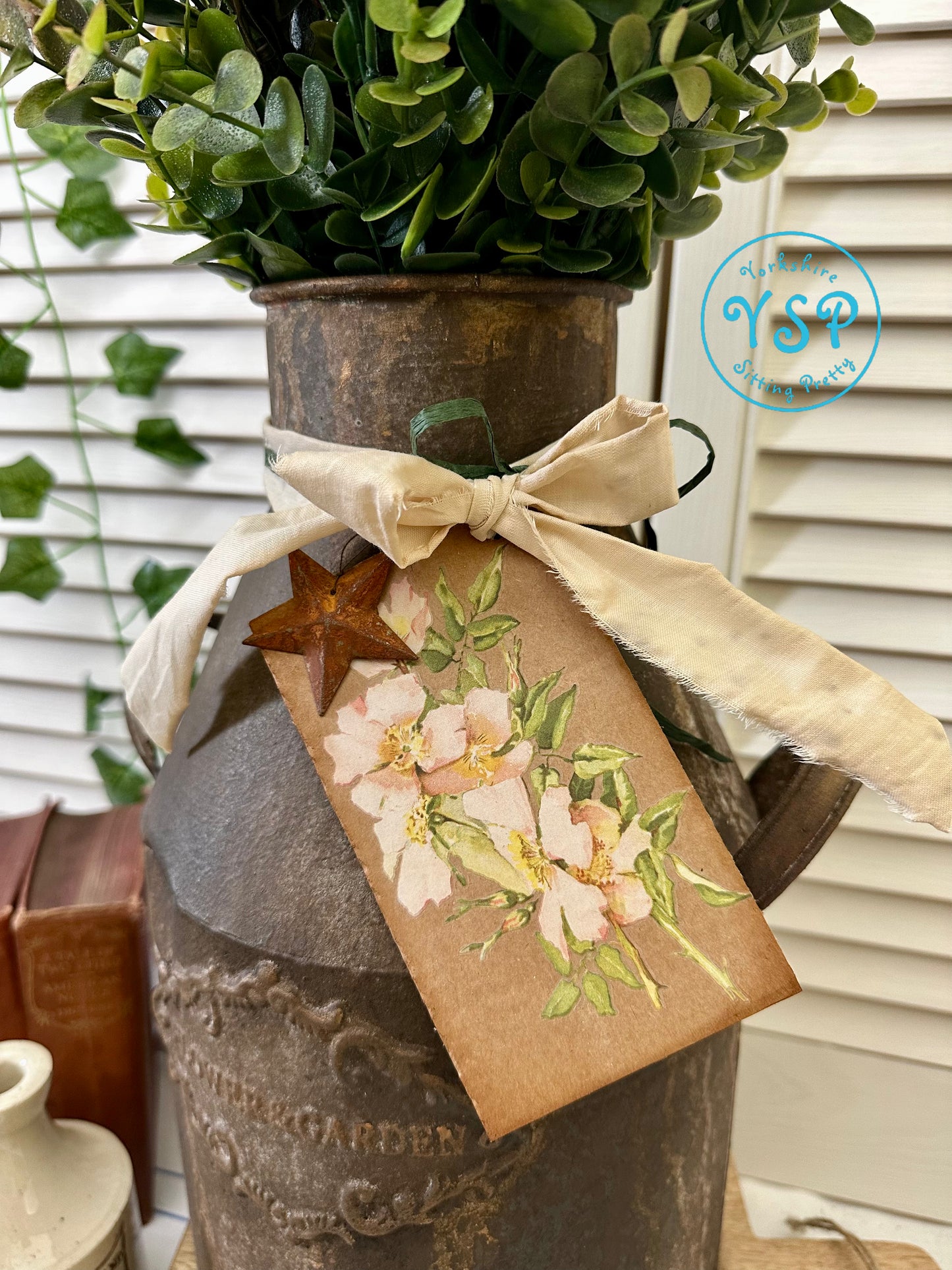Decorative rusty milk churn with a floral tag and bow, featuring a rustic design.