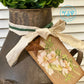 Decorative rusty milk churn with greenery, ribbon, and floral tag on a neutral background