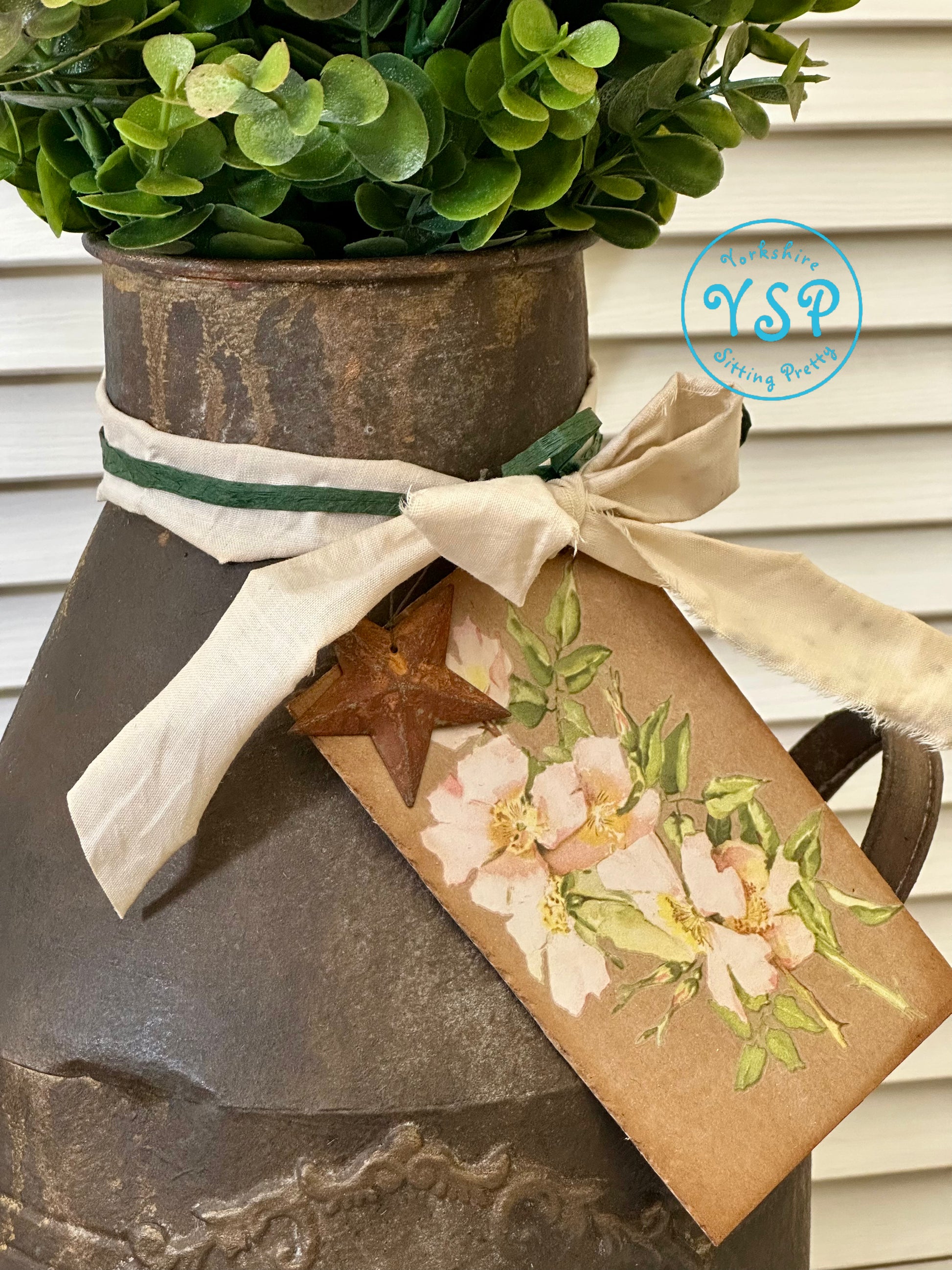 Decorative rusty milk churn with greenery, ribbon, and floral tag on a neutral background