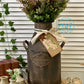 Decorative rusty milk churn with flowers and a tag on a white wall background