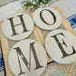Four round white coasters with 'HOME' letters on a textured surface with flowers and books.