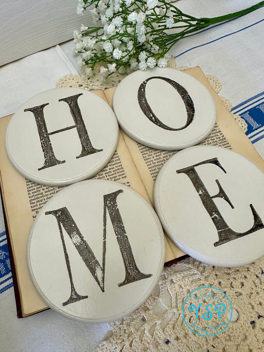 Four round white coasters with 'HOME' letters on a textured surface with flowers and books.