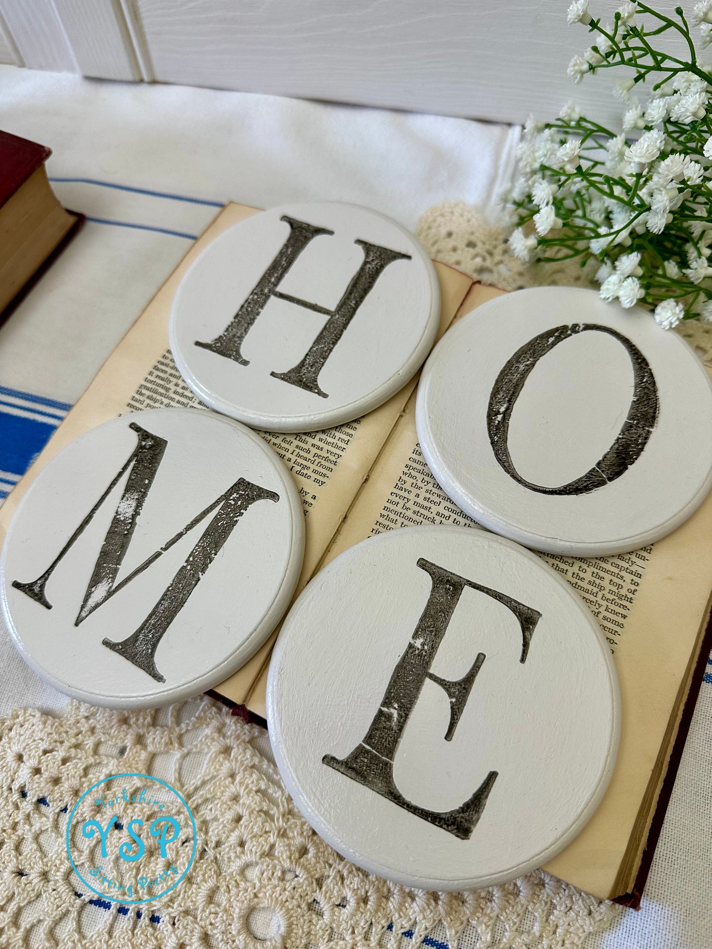 Four round white coasters with letters spelling 'HOME' on a open book background.