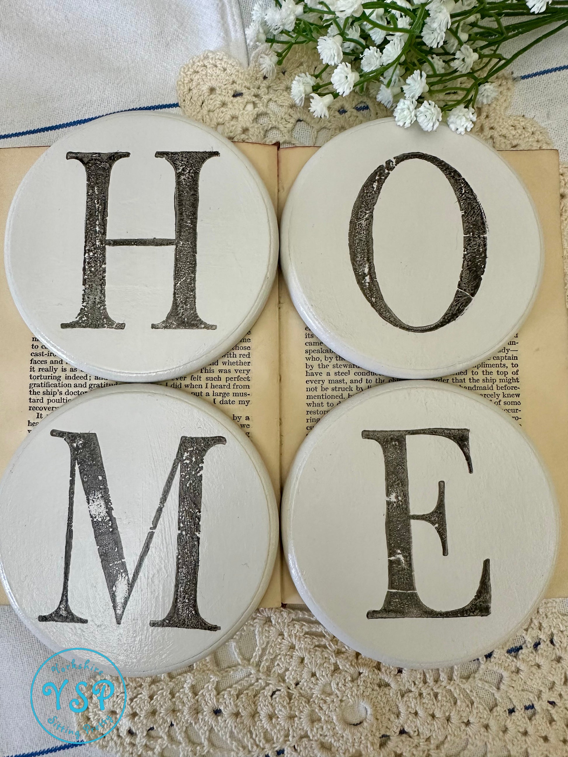 Decorative white round coasters spelling 'HOME' on a book with flowers and doily.