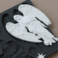 A white angel figurine with detailed wings and draped robes displayed on top of a dark gray decorative plaque featuring embossed holiday motifs and a small star piece, set against a solid taupe background. Silent Night mould from Iron Orchid Designs.