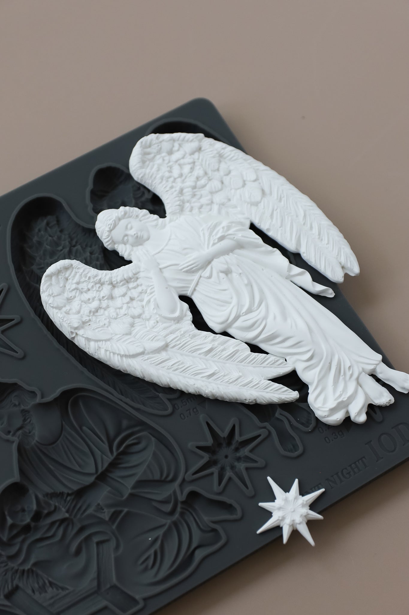 A white angel figurine with detailed wings and draped robes displayed on top of a dark gray decorative plaque featuring embossed holiday motifs and a small star piece, set against a solid taupe background. Silent Night mould from Iron Orchid Designs.