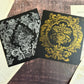 Two square black cards or boxes with ornate damask-style floral patterns, one in silver and one in gold, arranged on a rustic wooden surface with vintage book pages around them. Iron Orchid Designs
