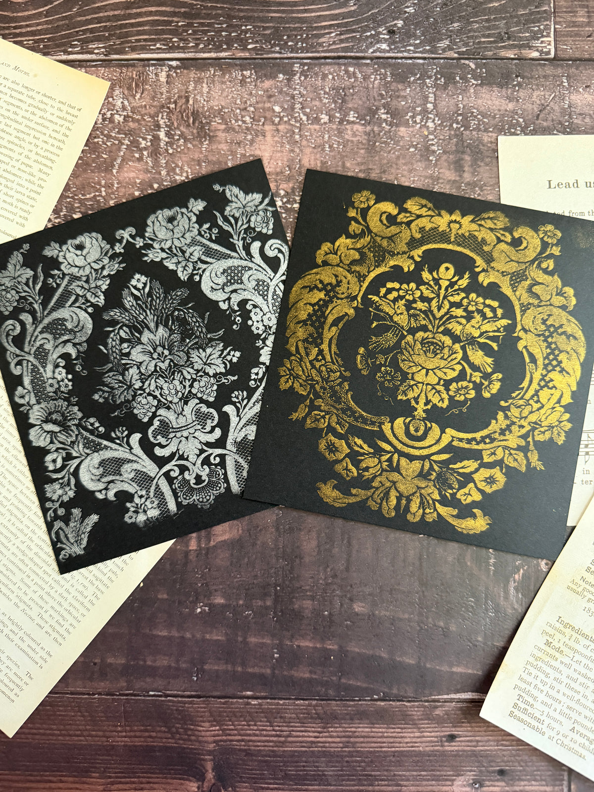Two square black cards or boxes with ornate damask-style floral patterns, one in silver and one in gold, arranged on a rustic wooden surface with vintage book pages around them. Iron Orchid Designs