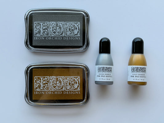 Two Iron Orchid Designs ink pads in dark gray and brown with silver and gold ink refills, shown with two small ink pad refill bottles on a light gray background.