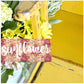 Yellow wooden sign with floral design and 'sunflower' text, surrounded by flowers.