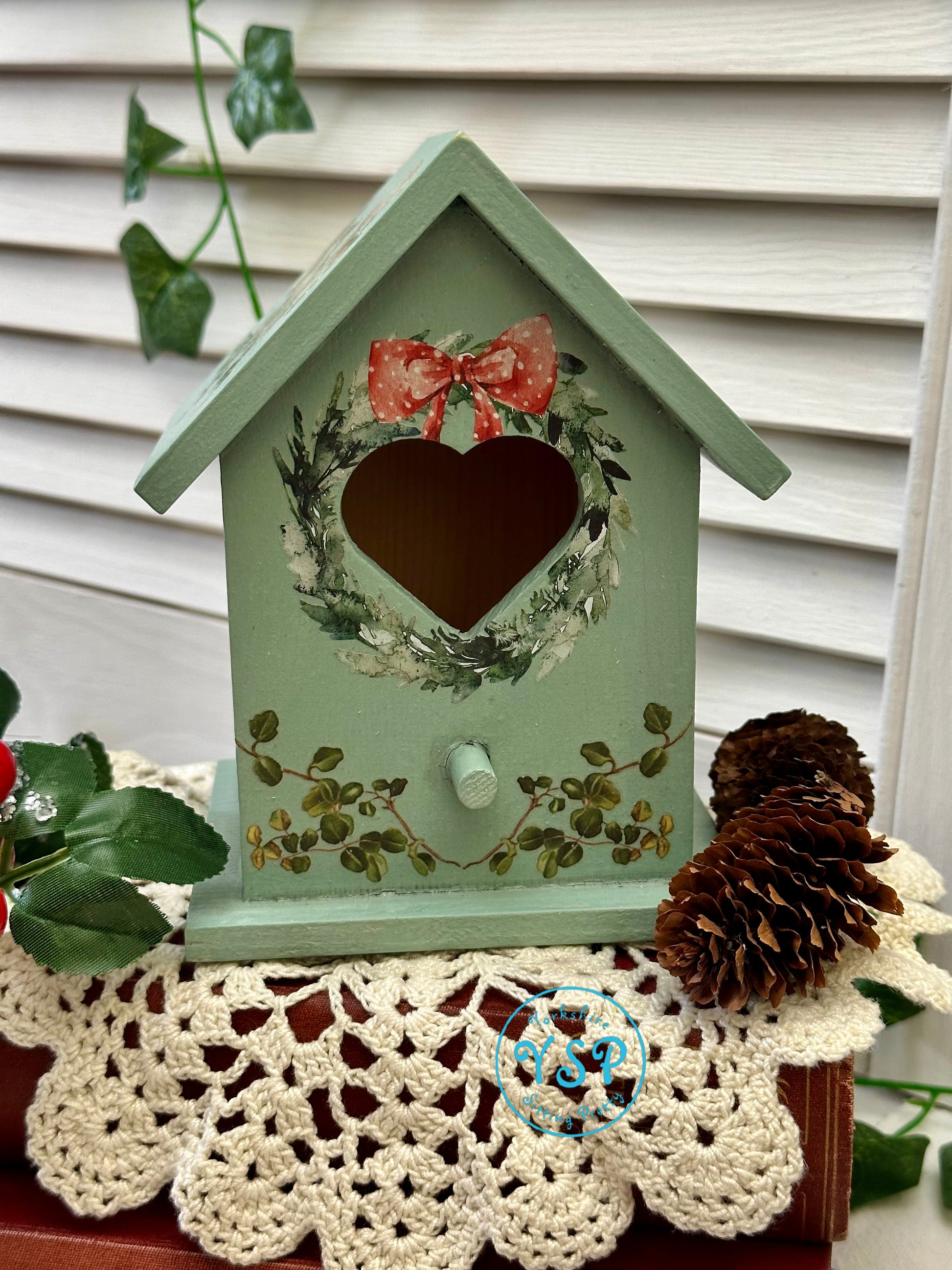 Decorative green birdhouse with a heart opening standing on a crotchet doily