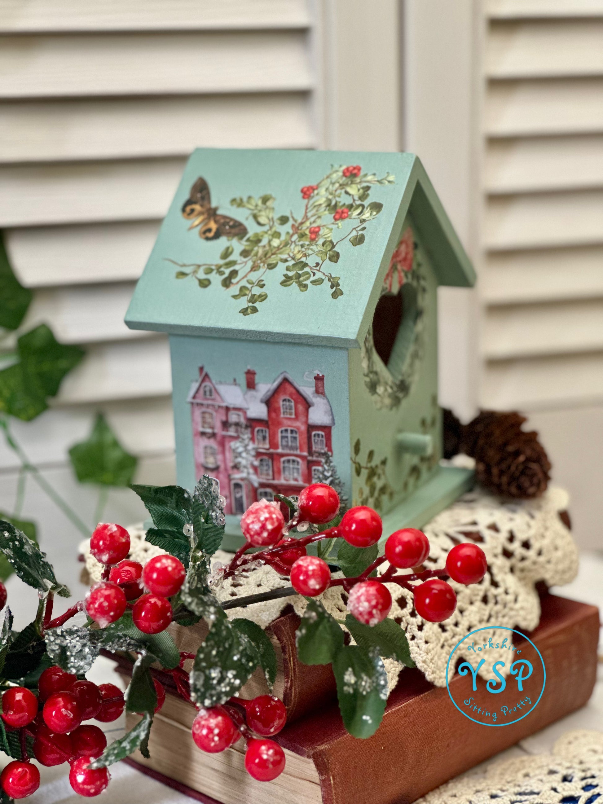 Decorative birdhouse with floral design on a stand with red berries and greenery.