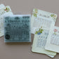 A set of monthly calendar cards with vintage floral designs and a central clear plastic box containing a stamp labeled 'A TIMELY TALE', arranged on a solid taupe background. A Timely Tale Stamp from Iron Orchid Designs.