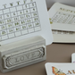 A tabletop desk calendar with a small white monthly calendar page in a decorative gray holder engraved with the word LOVE. Other calendar pages and printed cards are arranged around it on a beige surface. A Timely Tale Stamp from Iron Orchid Designs.