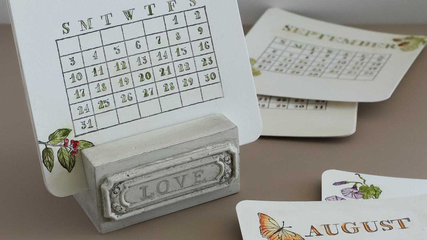 A tabletop desk calendar with a small white monthly calendar page in a decorative gray holder engraved with the word LOVE. Other calendar pages and printed cards are arranged around it on a beige surface. A Timely Tale Stamp from Iron Orchid Designs.