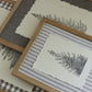 Three framed botanical art prints with light wood frames, layered diagonally. The prints feature delicate black stampings of fir trees on cream paper, set against backgrounds of linen in gray-brown, gray-and-white stripes, and brown-and-white gingham. Winter Grove stamp From Iron Orchid Designs.