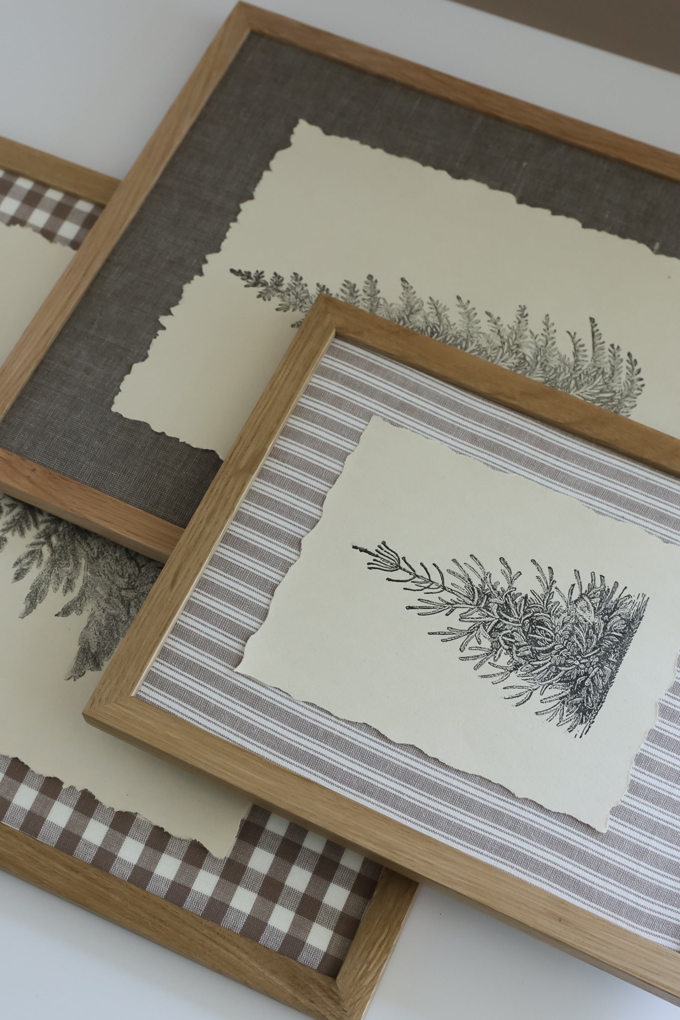 Three framed botanical art prints with light wood frames, layered diagonally. The prints feature delicate black stampings of fir trees on cream paper, set against backgrounds of linen in gray-brown, gray-and-white stripes, and brown-and-white gingham. Winter Grove stamp From Iron Orchid Designs.