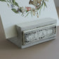 A small rectangular white and gray decorative stand with the word CREATE embossed on the front, holding a printed card with a floral illustration, on a beige surface. From iron Orchid Designs.