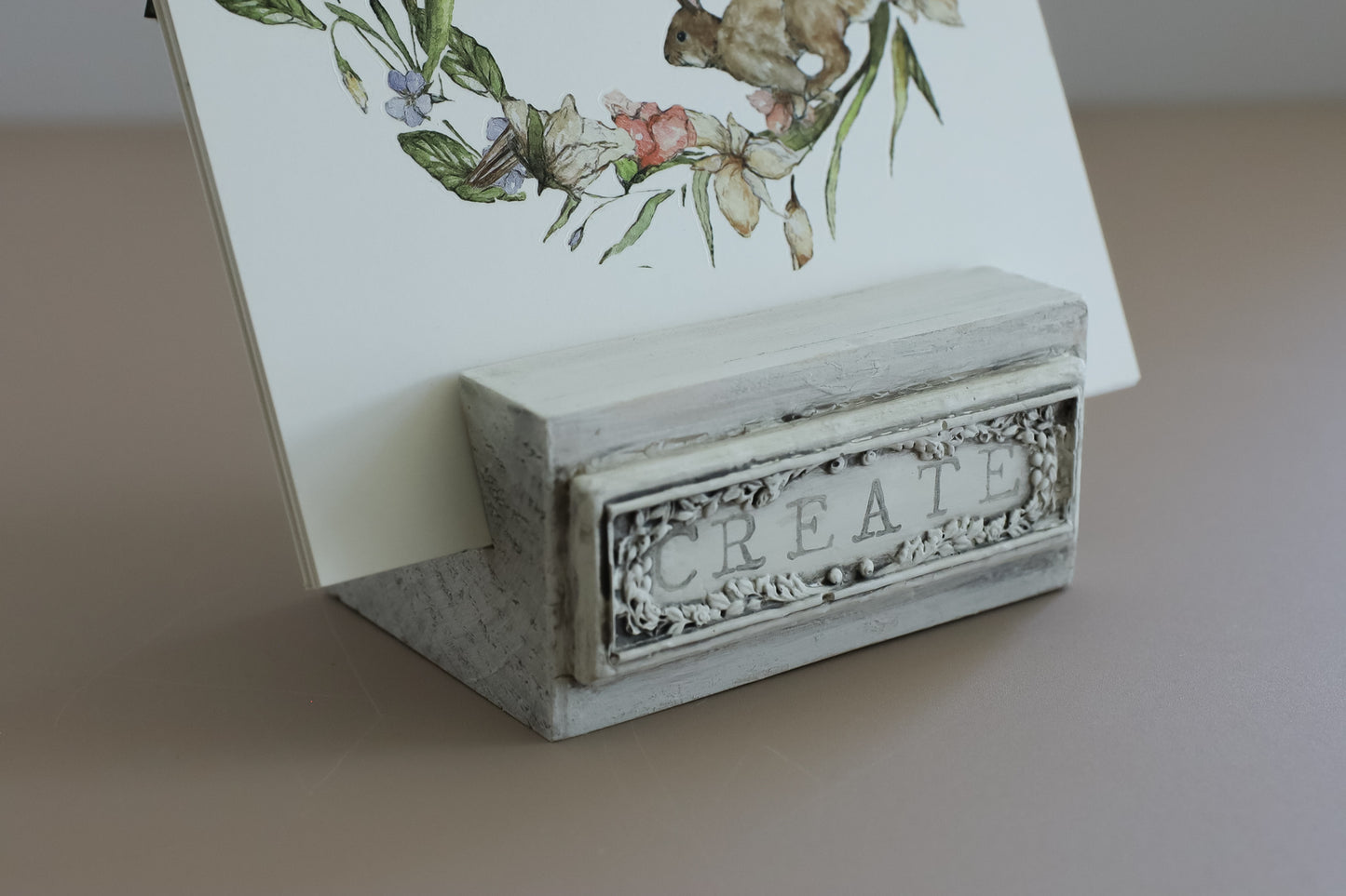 A small rectangular white and gray decorative stand with the word CREATE embossed on the front, holding a printed card with a floral illustration, on a beige surface. From iron Orchid Designs.