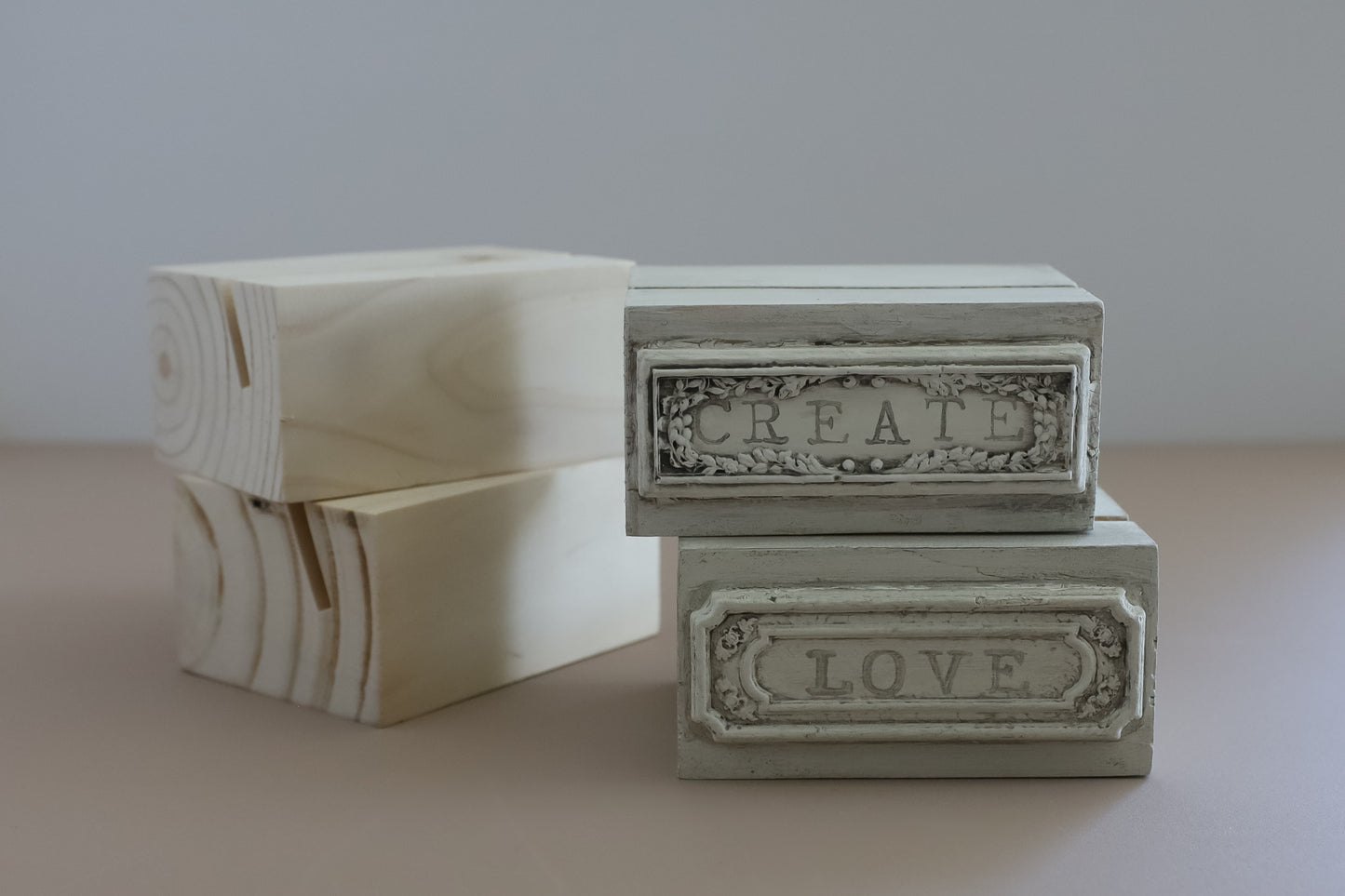 Four decorative rectangular wooden stands on a beige surface against a light gray background. Two boxes are unfinished light wood with visible grain, and two are painted in a distressed off-white finish with embossed labels reading CREATE and LOVE. From Iron Orchid Designs