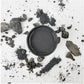 A black circular container filled with dark gray powder, surrounded by loose charcoal chunks and powder scattered on a white background. Paint Couture