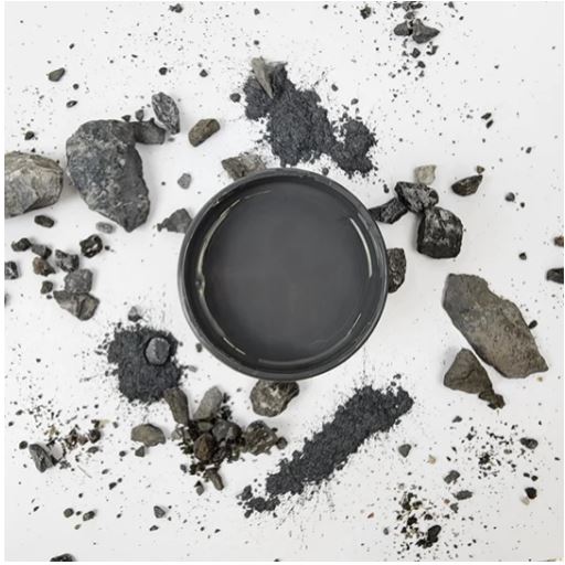 A black circular container filled with dark gray powder, surrounded by loose charcoal chunks and powder scattered on a white background. Paint Couture