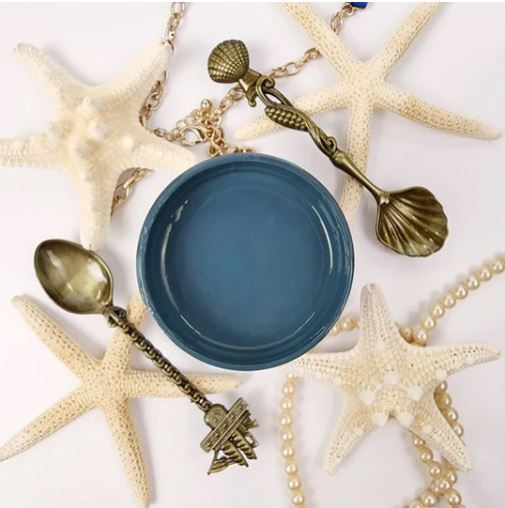 Pot of blue paint on a white cloth next to decorative spoons, beads and starfish. Paint Couture