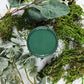 Pot of dark green paint surrounded by different leaves. Paint Couture