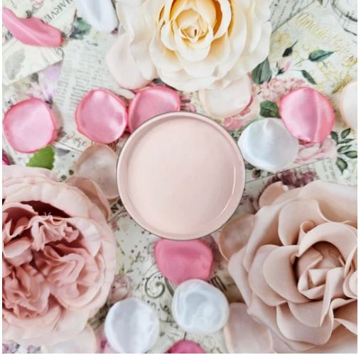 A round container filled with paint in a pale pink shade, surrounded by pink and cream roses, rose petals, and white petals on a vintage-style floral background. Paint Couture