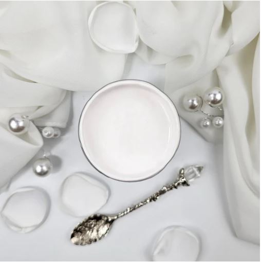 A pot of white paint, styled on a white fabric background with a silver spoon, pearl-like beads, and soft draped cloth. Paint Couture