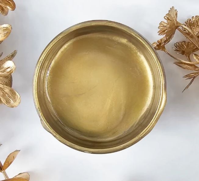 A round pot of Gold Lux Metallic from Paint Couture, photographed from directly above on a white background with decorative gold leaves in the corners.