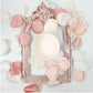 A pale peach paint in a small pot centred inside a painted decorative frame, surrounded by petals. Paint Couture