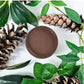A round pot of brown paint surrounded by green leaves and pinecones. Paint Couture