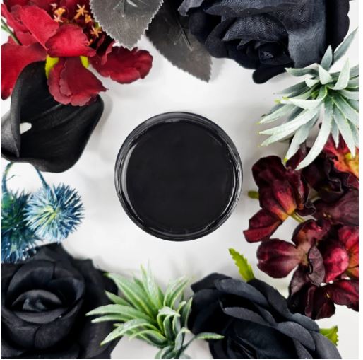 A round pot of black paint centered on a white background, surrounded by decorative black and dark red flowers and green foliage in a styled flat-lay arrangement. Paint Couture