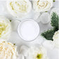 A small round pot of white paint, centered in a flat-lay arrangement of white and cream flowers and green leaves on a white background. Paint Couture