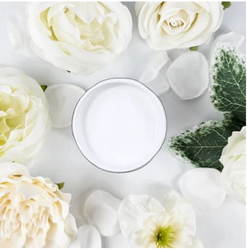 A small round pot of white paint, centered in a flat-lay arrangement of white and cream flowers and green leaves on a white background. Paint Couture