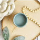 A small pot of blue paint standing on a log slice next to a string of wooden beads, leaves and a cream flower. Paint Couture
