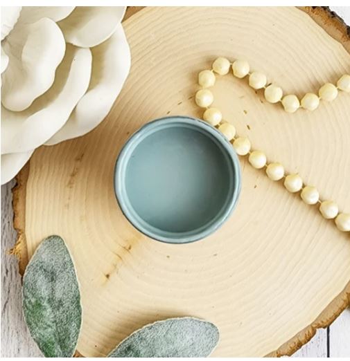 A small pot of blue paint standing on a log slice next to a string of wooden beads, leaves and a cream flower. Paint Couture