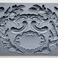 Iron Orchid Designs Olive Crest grey silicone mould on a white background. There are intricate designs of olive branches, swags, curls and a decorative label